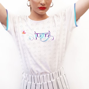 Puede incluir: Camiseta blanca con una pequeña mariposa y la palabra "Jugni" en escritura colorida. La camiseta tiene un ribete azul claro alrededor del cuello y las mangas.