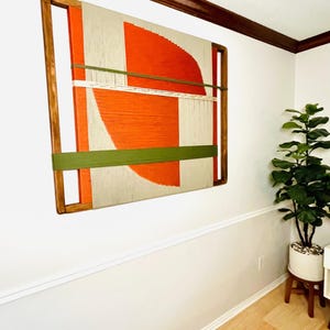a large abstract painting hanging on a white wall, with a wooden frame. The painting has a vibrant mix of orange and green colors, creating a striking visual contrast. The painting is positioned in the corner of the room, adding a unique decorative element to the space.