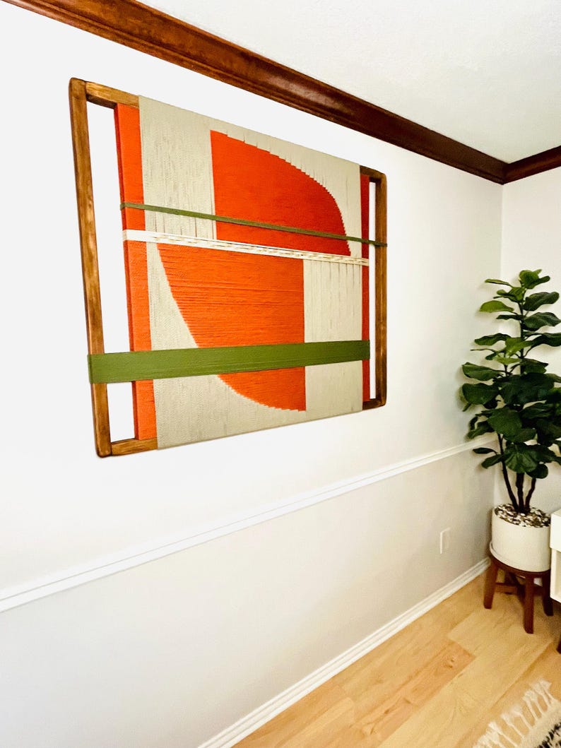 a large abstract painting hanging on a white wall, with a wooden frame. The painting has a vibrant mix of orange and green colors, creating a striking visual contrast. The painting is positioned in the corner of the room, adding a unique decorative element to the space.