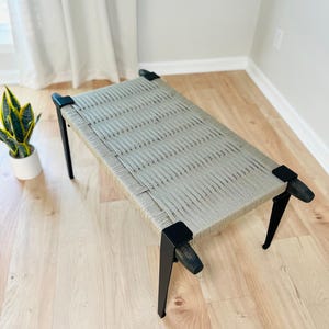 May include: A rectangular bench featuring a woven light gray cord seat stretched across a black wooden frame. The bench has four tapered black legs with square corner joints that secure the woven cord surface in a tight, linear pattern.