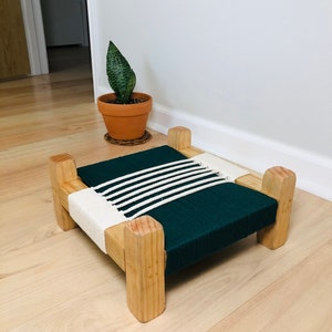 May include: A small wooden pet bed with a dark green fabric base and white rope accents. The bed is sitting on a light wood floor.