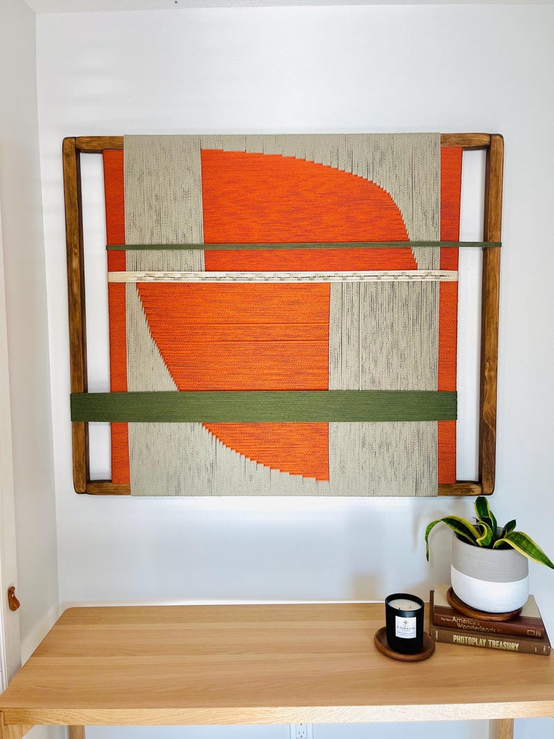 a wooden table with a plant and a candle on it, set against a white wall with a large abstract painting hanging above.