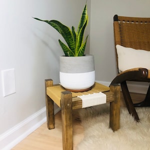May include: A potted snake plant on a wooden plant stand with a woven top. The stand is in a corner of a room with a white wall and a light brown wood floor.