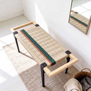May include: A rectangular bench featuring a light-colored wooden frame with black metal legs and corner brackets. The seat is constructed from woven beige cord, accented by a single prominent stripe in dark green and a thinner stripe in burnt orange.