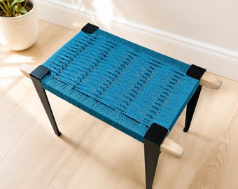 May include: A rectangular stool featuring a woven teal cord seat stretched across a light wood frame. The stool has four tapered black legs with matching black corner brackets that secure the woven seat to the wooden support structure.