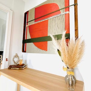 a wooden desk with a vase of dried flowers, a stack of books, and a large abstract painting hanging on the wall above the desk.