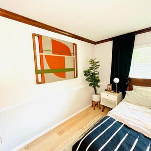 a bedroom with a bed, a nightstand, and a large abstract painting on the wall. The room has a white color scheme and features a rug on the floor.