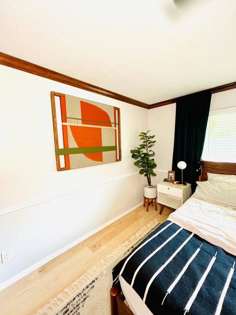 a bedroom with a bed, a nightstand, and a large abstract painting on the wall. The room has a white color scheme and features a rug on the floor.