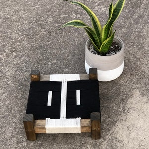 May include: A black and white woven stool with a wooden frame. The stool is sitting on a concrete surface next to a potted snake plant.