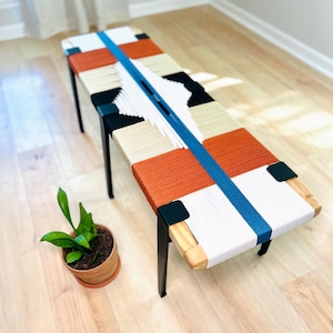 May include: A woven bench with a geometric pattern in orange, white, and black yarn. The bench has black metal legs and a wooden frame.