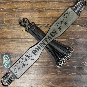 May include: A gray leather duck call lanyard featuring a wide neck strap engraved with the name FOUNTAIN and a flock of flying ducks. The strap includes black woven ends, metal swivel clips, and multiple black drop loops with silver metal rings.