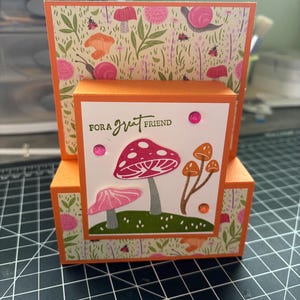 May include: A handmade greeting card with an orange base and a whimsical mushroom design. The card features the text "FOR A great FRIEND" and is decorated with pink and orange mushrooms, green grass, and pink embellishments. The background has a floral pattern.