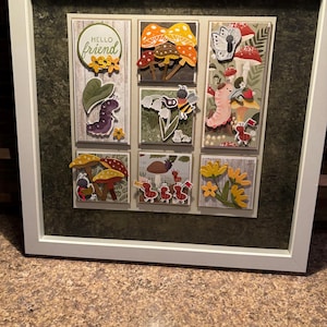 May include: A framed artwork featuring nine small, illustrated scenes. The scenes depict whimsical mushrooms, caterpillars, butterflies, and flowers. The top left scene has the text "Hello Friend" in a circle. The frame is white.