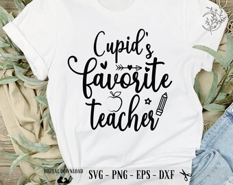 以下が含まれることがあります： 黒い文字で「Cupid's favorite teacher」と書かれた白いTシャツ。小さなリンゴ、矢印、鉛筆のグラフィックが描かれている。