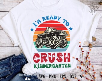 Puede incluir: Camiseta blanca con un gráfico colorido que presenta un camión monstruo negro y el texto "I'M READY TO CRUSH KINDERGARTEN." El diseño incluye un atardecer rojo y naranja, estrellas azules y texto verde.