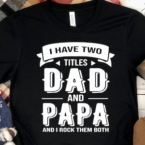 Pode incluir: Camiseta preta com texto branco que diz "Eu tenho dois títulos Pai e Papá e eu os rockeo ambos".