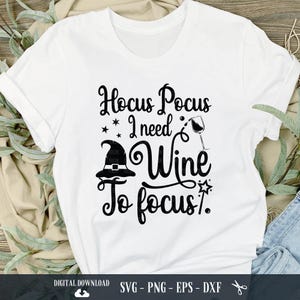 Op de afbeelding: Wit T-shirt met zwarte tekst die "Hocus Pocus I need Wine To Focus." luidt. Een heksenhoed en een glas wijn zijn opgenomen in het ontwerp.