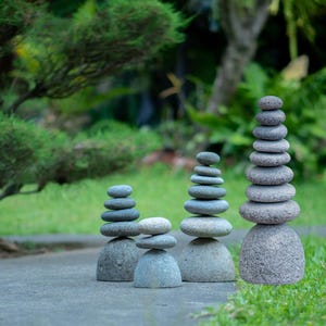 May include: Four stacks of gray stones of varying heights are balanced on a concrete surface, with a blurred green background of grass and foliage. The stacks range from three to ten stones each, creating a sense of balance and tranquility.