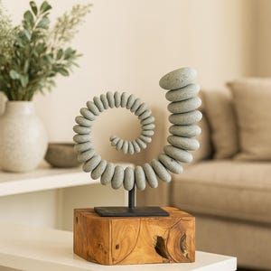 May include: A decorative sculpture featuring a spiral design made of gray stones. The stones are stacked on a black metal stand, which is mounted on a wooden block. The sculpture is a unique home decor item.