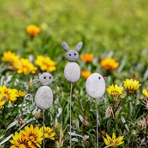 May include: Three whimsical garden stakes featuring stone animal figures: a cat with wire whiskers, a bunny with upright ears, and a bird. The figures are gray and set against a backdrop of green grass and yellow flowers, creating a charming outdoor scene.