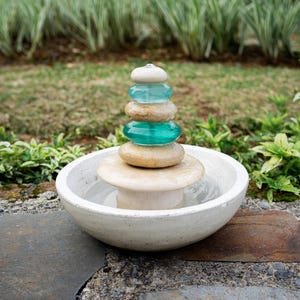 Hand-carved white marble fountain with luminous natural stone base and stacked cairns made from polished jasper and translucent recycled blue glass. Water flows gently over each tier, creating a soothing rippling sound and serene visual effect. A heavy, durable tabletop fountain perfect for meditation, yoga spaces, spa-inspired décor, or as a calming centerpiece for living rooms, covered patios, or garden nooks. Unique handmade design with clean cord-free look.