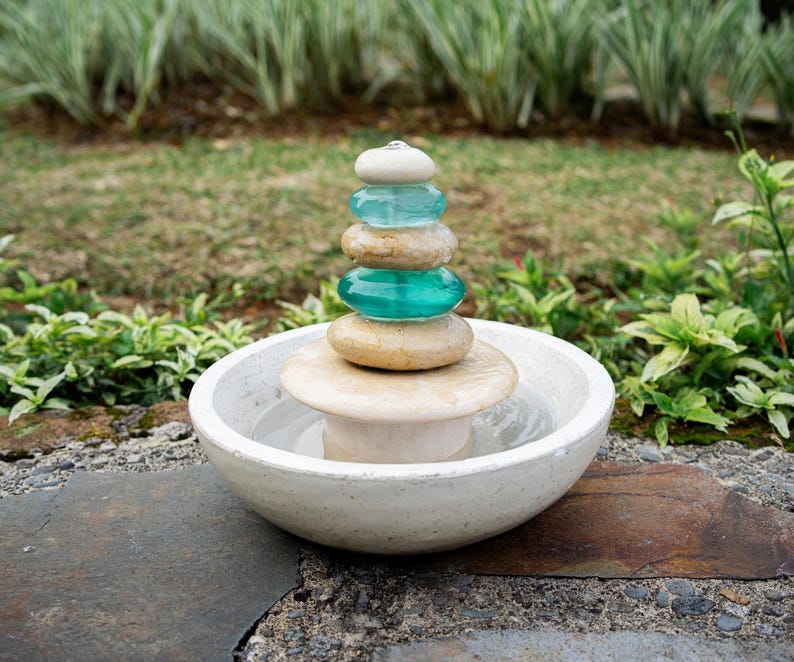 Hand-carved white marble fountain with luminous natural stone base and stacked cairns made from polished jasper and translucent recycled blue glass. Water flows gently over each tier, creating a soothing rippling sound and serene visual effect. A heavy, durable tabletop fountain perfect for meditation, yoga spaces, spa-inspired décor, or as a calming centerpiece for living rooms, covered patios, or garden nooks. Unique handmade design with clean cord-free look.