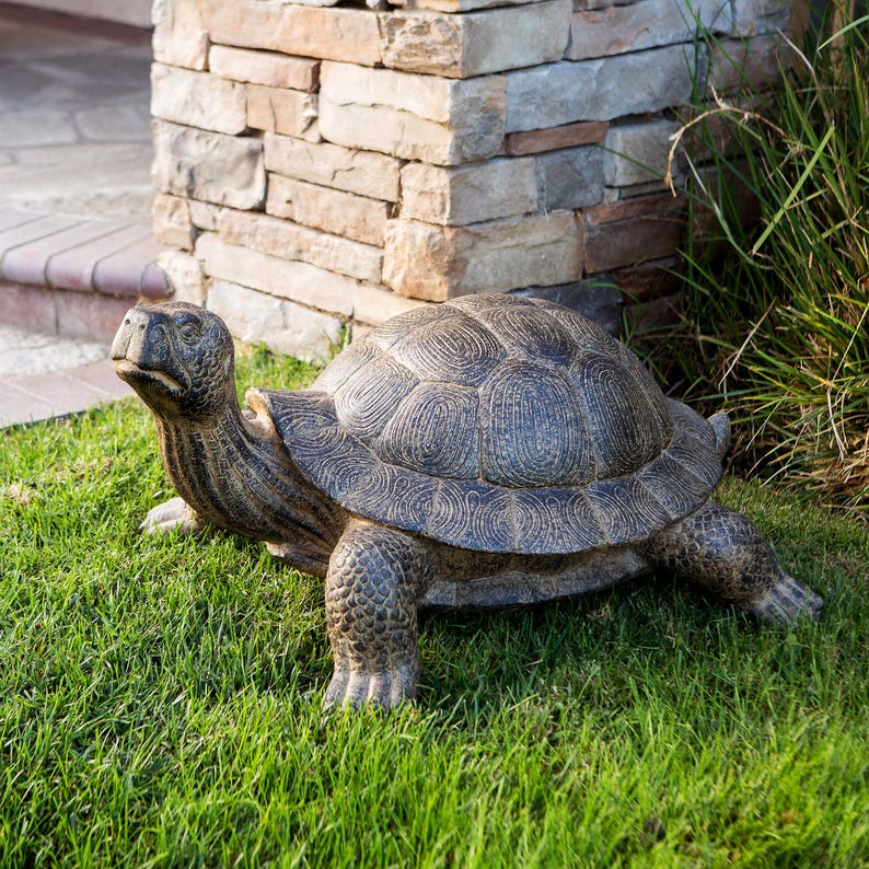 May include: A large, detailed turtle statue in shades of gray and brown rests on a bed of green grass. The statue has a textured shell and detailed legs and head. The turtle is positioned near a stone pillar.