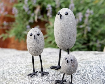 May include: Three stone bird sculptures with black metal legs and beaks. The birds are made of gray stones with black eyes. The sculptures are of varying sizes, with the largest bird in the center.