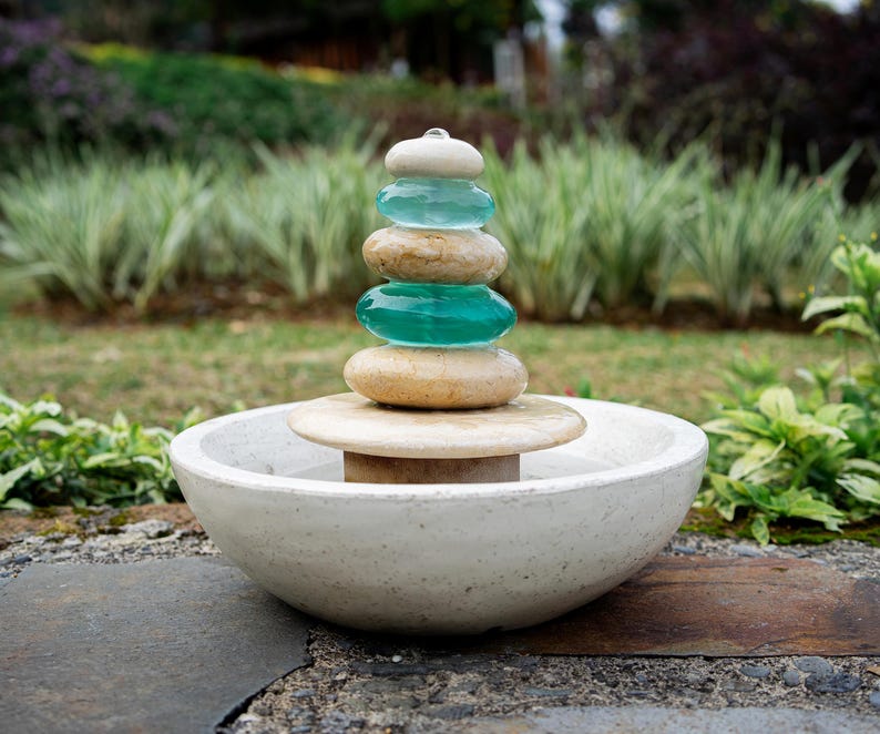 Hand-carved white marble fountain with luminous natural stone base and stacked cairns made from polished jasper and translucent recycled blue glass. Water flows gently over each tier, creating a soothing rippling sound and serene visual effect. A heavy, durable tabletop fountain perfect for meditation, yoga spaces, spa-inspired décor, or as a calming centerpiece for living rooms, covered patios, or garden nooks. Unique handmade design with clean cord-free look.