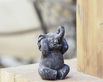 May include: A small, gray elephant figurine in a seated pose, with its trunk raised and ears covering its eyes. The statue has a weathered appearance, suggesting an outdoor garden ornament. The elephant is sitting on a brick surface.