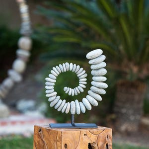 May include: A stone spiral sculpture, crafted from smooth, gray river rocks, is mounted on a black metal stand. The sculpture is displayed on a natural wood block, with a blurred green background of foliage.
