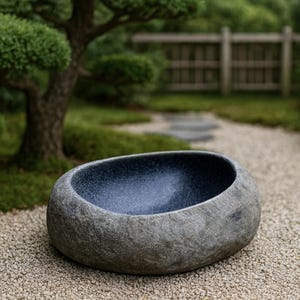 Large 13–14 inch hand-carved river stone bird bath placed in a peaceful garden setting with gravel ground and manicured shrubs. The oversized bowl features a smooth, dark polished interior and a thick, natural stone exterior with organic gray texture. Its substantial weight and rounded shape make it ideal as a focal point in zen gardens, patios, and outdoor landscapes, inviting birds to drink and bathe.
