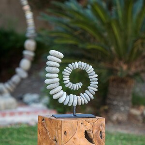 May include: A spiral sculpture made of small, gray stones, mounted on a black metal stand. The sculpture sits atop a square, wooden block. The background features green foliage and a blurred view of a garden setting.