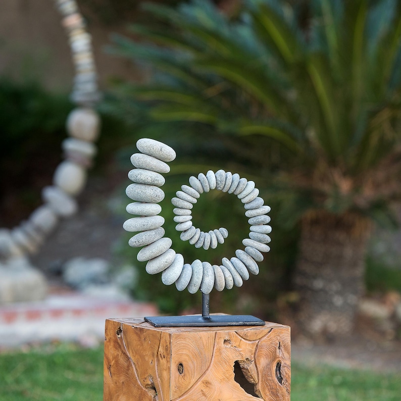 May include: A spiral sculpture made of small, gray stones, mounted on a black metal stand. The sculpture sits atop a square, wooden block. The background features green foliage and a blurred view of a garden setting.