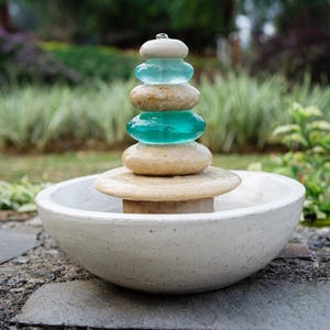 Hand-carved white marble fountain with luminous natural stone base and stacked cairns made from polished jasper and translucent recycled blue glass. Water flows gently over each tier, creating a soothing rippling sound and serene visual effect. A heavy, durable tabletop fountain perfect for meditation, yoga spaces, spa-inspired décor, or as a calming centerpiece for living rooms, covered patios, or garden nooks. Unique handmade design with clean cord-free look.