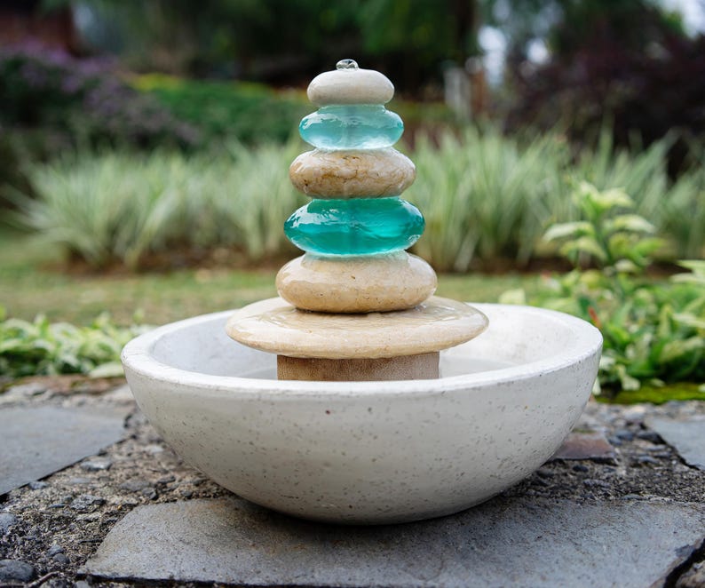 Hand-carved white marble fountain with luminous natural stone base and stacked cairns made from polished jasper and translucent recycled blue glass. Water flows gently over each tier, creating a soothing rippling sound and serene visual effect. A heavy, durable tabletop fountain perfect for meditation, yoga spaces, spa-inspired décor, or as a calming centerpiece for living rooms, covered patios, or garden nooks. Unique handmade design with clean cord-free look.