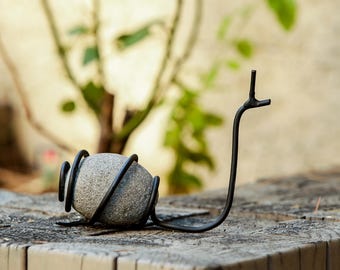 May include: A decorative metal snail sculpture. The snail's body is a gray stone, encircled by a black metal coil. The snail's head and antennae are crafted from black metal, creating a whimsical garden accent.