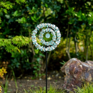 May include: A decorative garden stake featuring a spiral design. The spiral is made of light blue and white stones and wooden beads. A light blue glass orb hangs in the center. The stake is black and is set against a blurred green background.