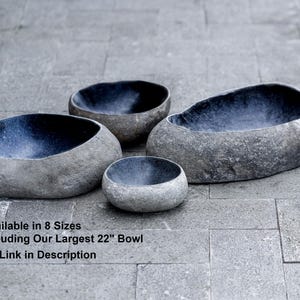 Multiple river stone bird bath bowls displayed together outdoors, showing a full range of sizes from small to extra-large. Each bowl features a hand-carved natural stone exterior with a smooth, dark polished interior. The group photo highlights variations in shape, thickness, and color across the collection. Perfect for showcasing all available sizes and the organic, heavy, durable nature of genuine river stone garden bowls.