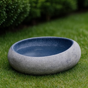 Large 13–14 inch hand-carved river stone bird bath placed in a peaceful garden setting with gravel ground and manicured shrubs. The oversized bowl features a smooth, dark polished interior and a thick, natural stone exterior with organic gray texture. Its substantial weight and rounded shape make it ideal as a focal point in zen gardens, patios, and outdoor landscapes, inviting birds to drink and bathe.