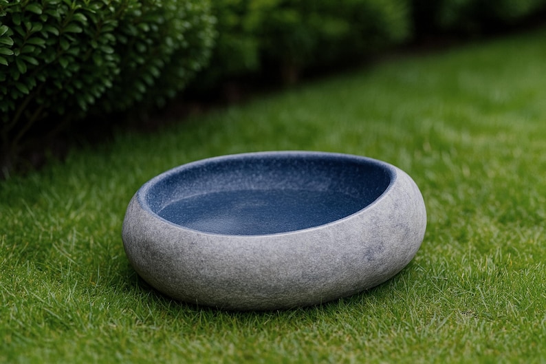 Large 13–14 inch hand-carved river stone bird bath placed in a peaceful garden setting with gravel ground and manicured shrubs. The oversized bowl features a smooth, dark polished interior and a thick, natural stone exterior with organic gray texture. Its substantial weight and rounded shape make it ideal as a focal point in zen gardens, patios, and outdoor landscapes, inviting birds to drink and bathe.