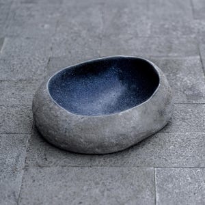 Large 13–14 inch hand-carved river stone bird bath placed in a peaceful garden setting with gravel ground and manicured shrubs. The oversized bowl features a smooth, dark polished interior and a thick, natural stone exterior with organic gray texture. Its substantial weight and rounded shape make it ideal as a focal point in zen gardens, patios, and outdoor landscapes, inviting birds to drink and bathe.
