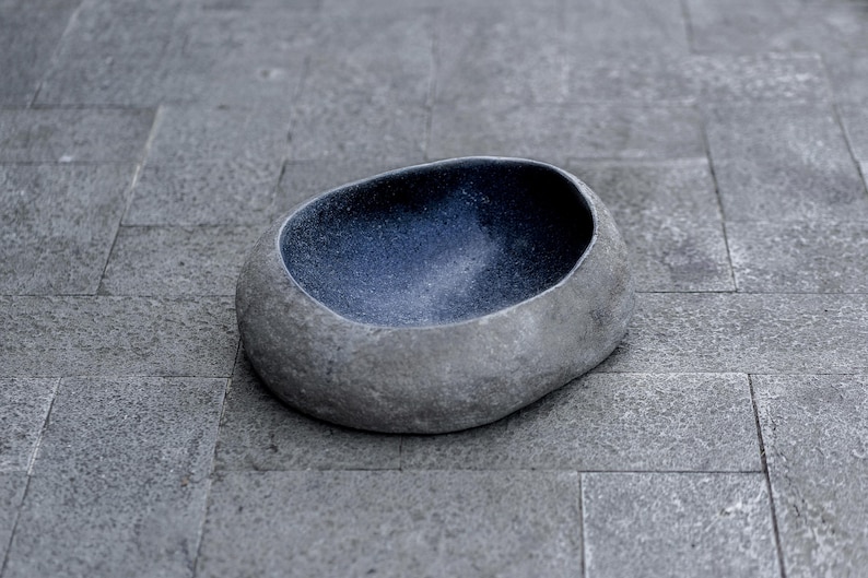Large 13–14 inch hand-carved river stone bird bath placed in a peaceful garden setting with gravel ground and manicured shrubs. The oversized bowl features a smooth, dark polished interior and a thick, natural stone exterior with organic gray texture. Its substantial weight and rounded shape make it ideal as a focal point in zen gardens, patios, and outdoor landscapes, inviting birds to drink and bathe.