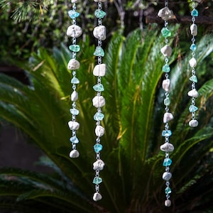May include: Decorative hanging chains with alternating natural stones and blue-green glass beads. The chains are suspended against a backdrop of green foliage, creating a natural and organic aesthetic. The stones are a light gray color.