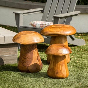 May include: Three wooden mushroom-shaped stools of varying sizes are displayed on a green lawn. The stools are made of light brown wood with a natural grain pattern. A gray wooden chair is in the background.