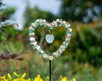 May include: A heart-shaped garden stake with a decorative design. The heart is crafted from small, light blue and wood-colored pieces, with a small, pale blue heart pendant hanging in the center. The stake is black and set in a garden.