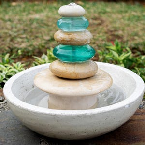 Hand-carved white marble fountain with luminous natural stone base and stacked cairns made from polished jasper and translucent recycled blue glass. Water flows gently over each tier, creating a soothing rippling sound and serene visual effect. A heavy, durable tabletop fountain perfect for meditation, yoga spaces, spa-inspired décor, or as a calming centerpiece for living rooms, covered patios, or garden nooks. Unique handmade design with clean cord-free look.