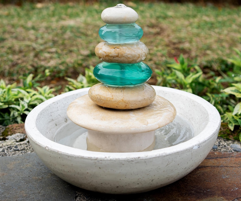Hand-carved white marble fountain with luminous natural stone base and stacked cairns made from polished jasper and translucent recycled blue glass. Water flows gently over each tier, creating a soothing rippling sound and serene visual effect. A heavy, durable tabletop fountain perfect for meditation, yoga spaces, spa-inspired décor, or as a calming centerpiece for living rooms, covered patios, or garden nooks. Unique handmade design with clean cord-free look.