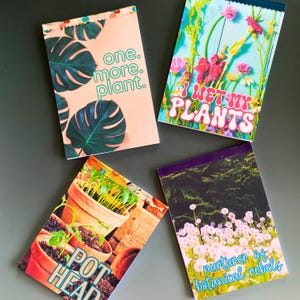 May include: Four rectangular notebooks with botanical themes. One features monstera leaves and the text 'one. more. plant.' Another shows colourful flowers with the text 'I WET MY PLANTS'. A third displays potted plants with 'POT HEAD' text. The last shows dandelions with 'nurturer of botanical rebels' text.