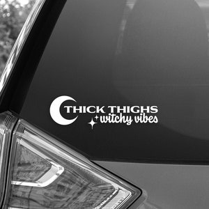 May include: A black car with a white vinyl sticker on the back window. The sticker says "Thick Thighs + Witchy Vibes" with a crescent moon and a star.
